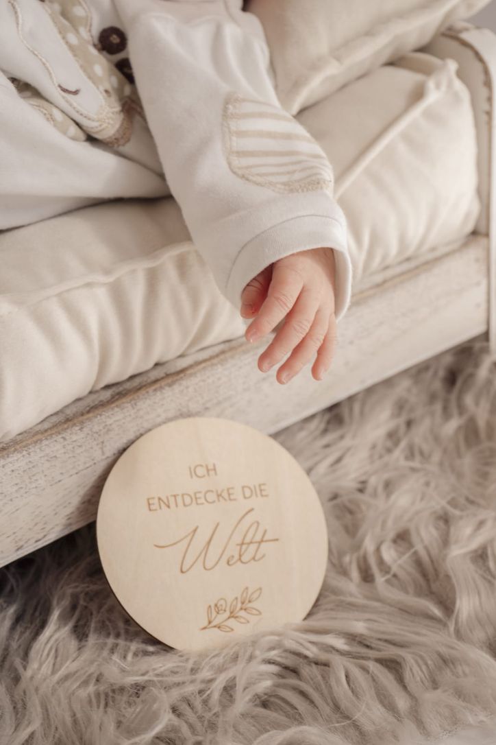 Nahaufnahme der Hand eines Neugeborenen neben Holzschild „Ich entdecke die Welt"