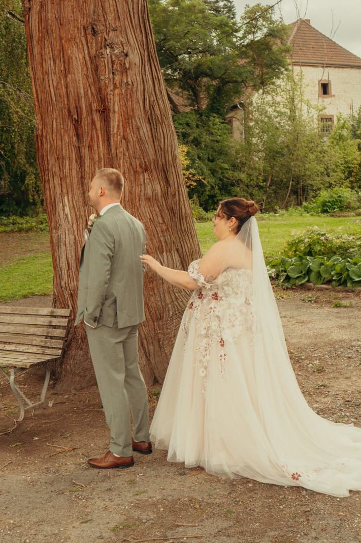 Braut berührt sanft die Schulter ihres Bräutigams beim First Look unter einem großen Baum