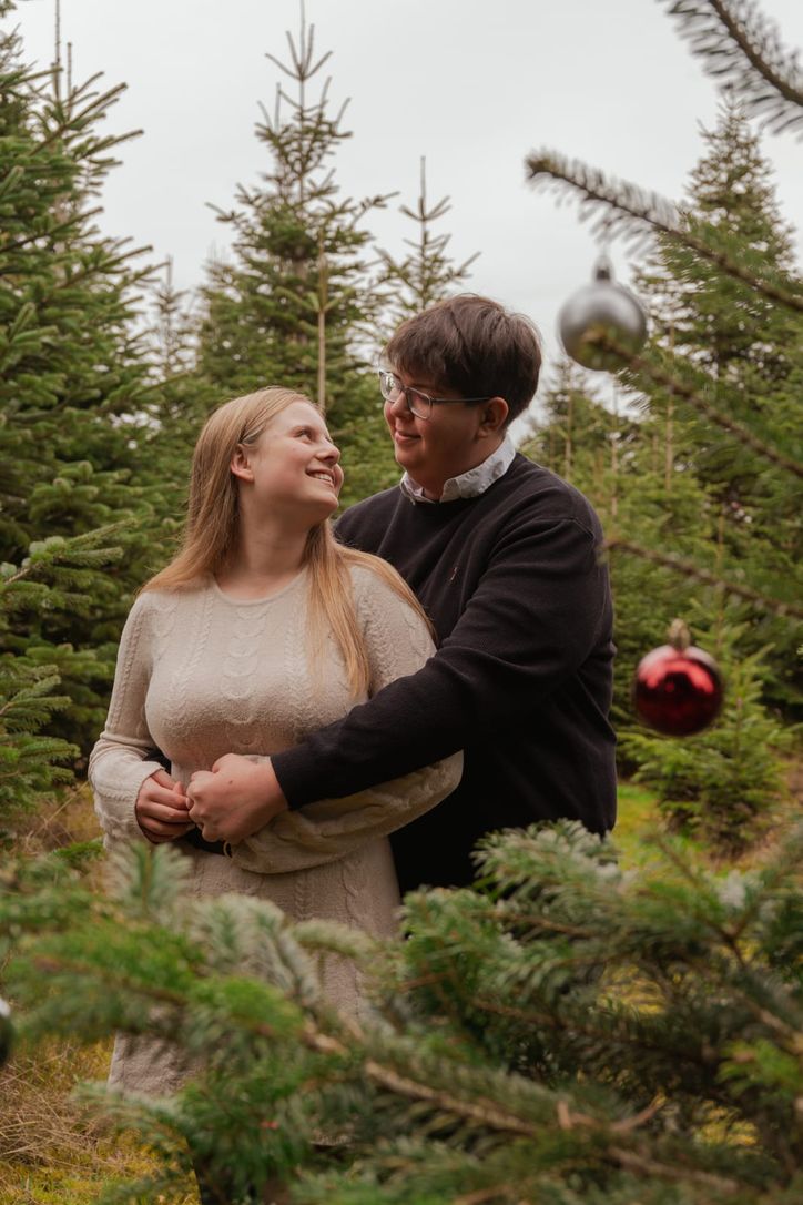 Paar umarmt sich lächelnd zwischen Christbäumen mit hängenden Kugeln