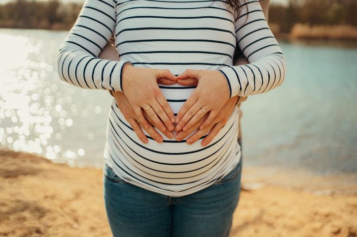 Schwangere formt mit ihren Händen ein Herz auf dem Babybauch am See
