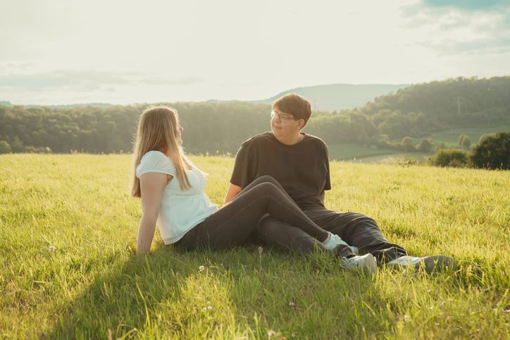 Paar sitzt lachend im Gras vor weitem Blick auf hügelige Landschaft im Abendlicht